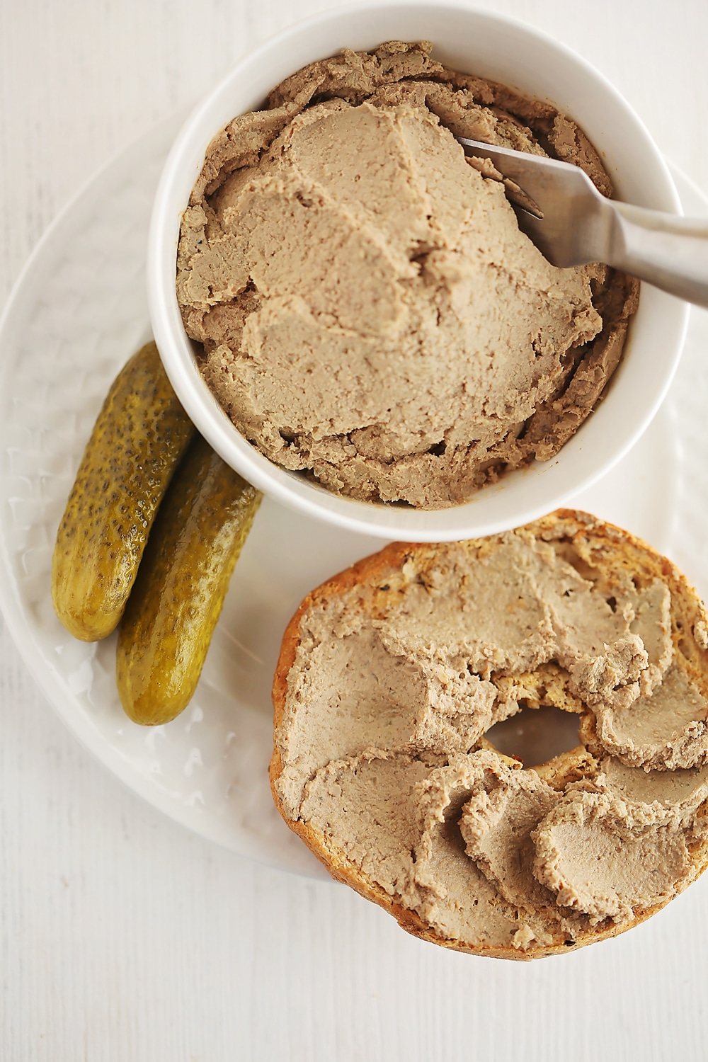 liver pate spread on top of the bagel with pickle next to it, and small white bowl filled with pate