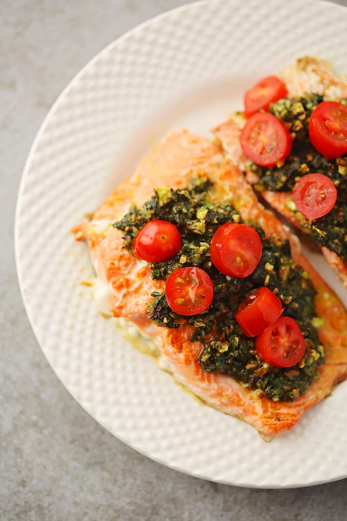 white plate with cooked salmon slice topped with green sauce and bright red grape tomatoes