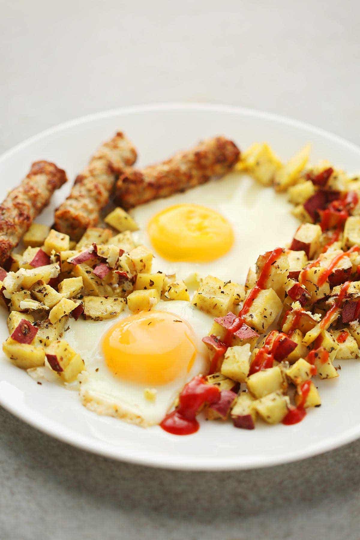 white plate with sunny side up eggs, cubed potatoes with ketchup sauce