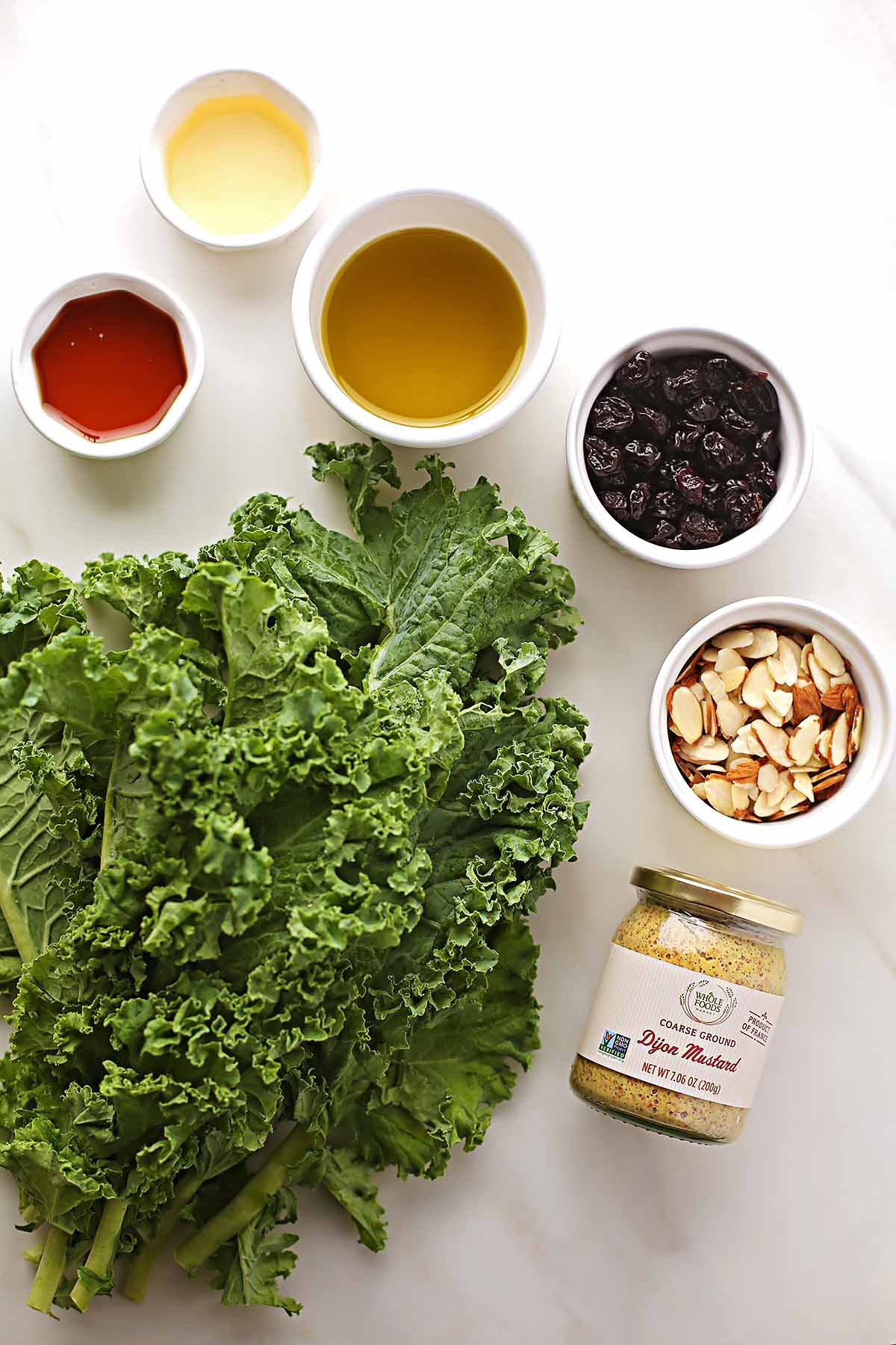 tabletop with prepared ingredients to make salad with kale: kale, white bowls with dried berries, almonds, dressing