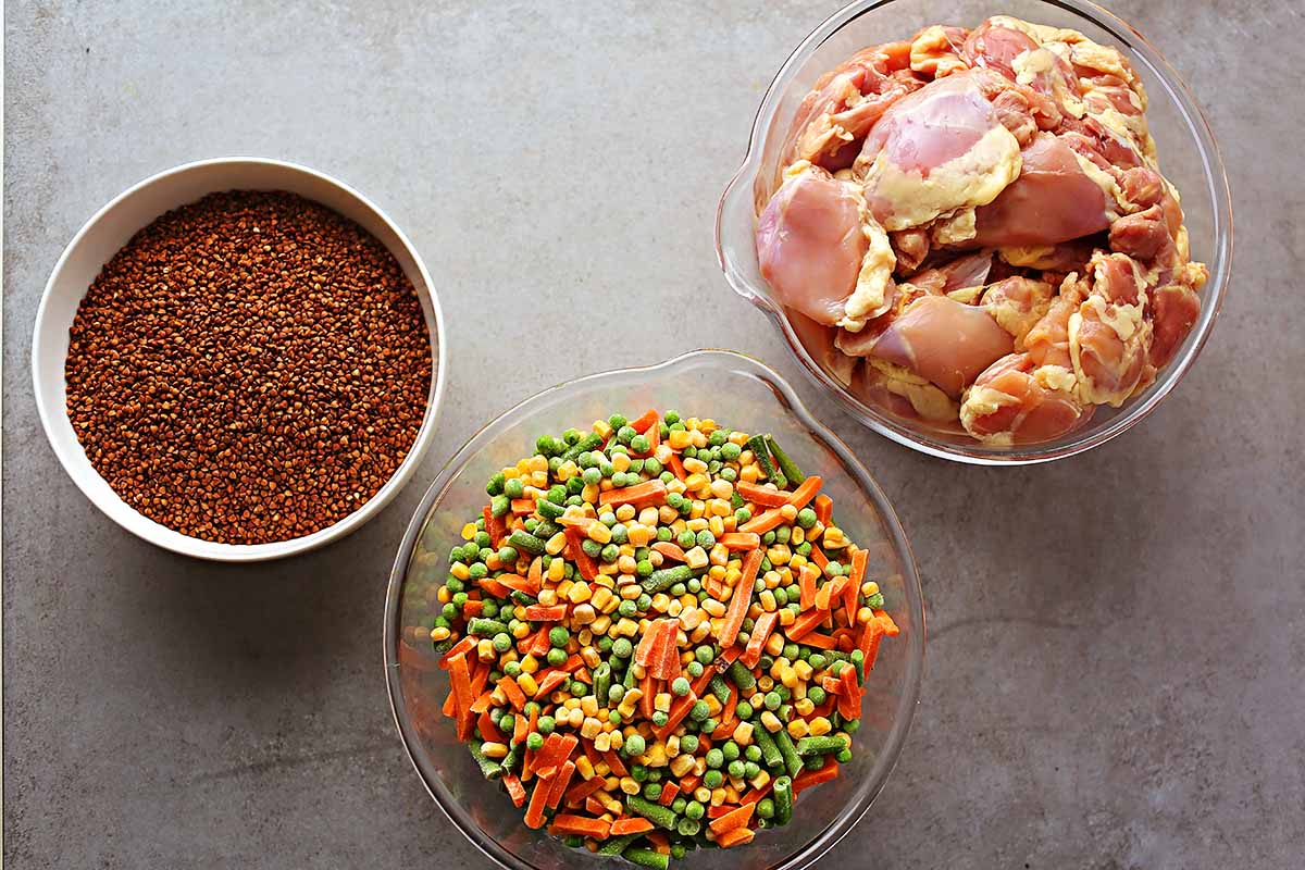 tabletop with ingredients for making dog food: uncooked buckwheat groats, frozen mixed vegetables, raw chicken meat