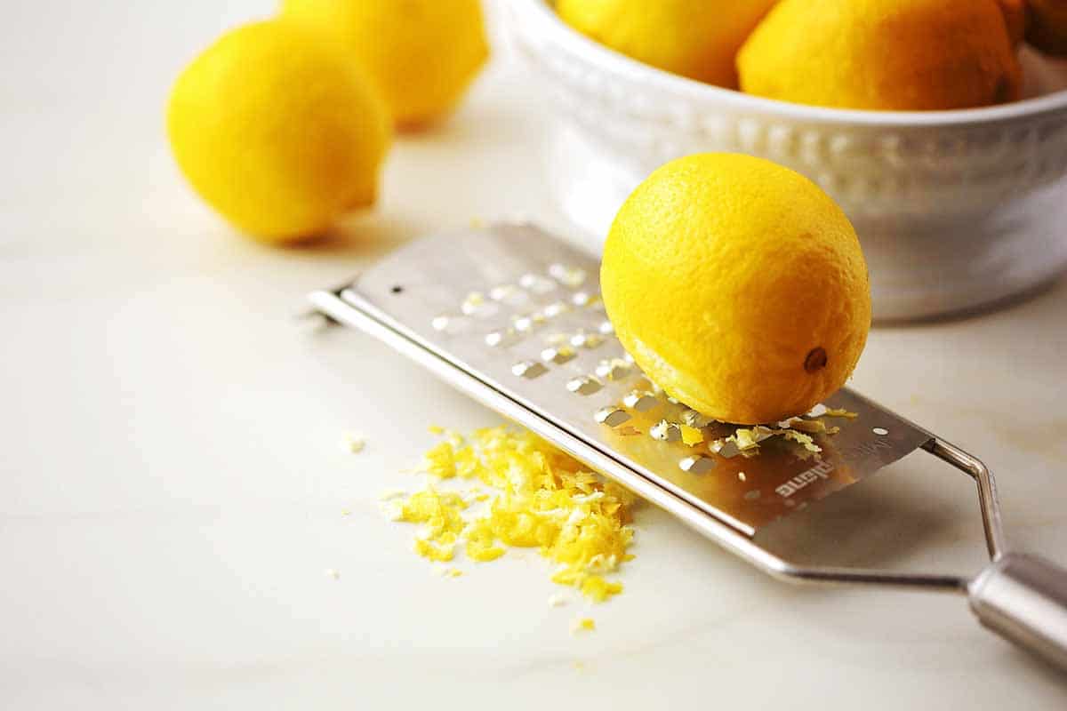 tabletop with stainless steel lemon zester, lemon shreds, whole lemon, white bowl with lemons