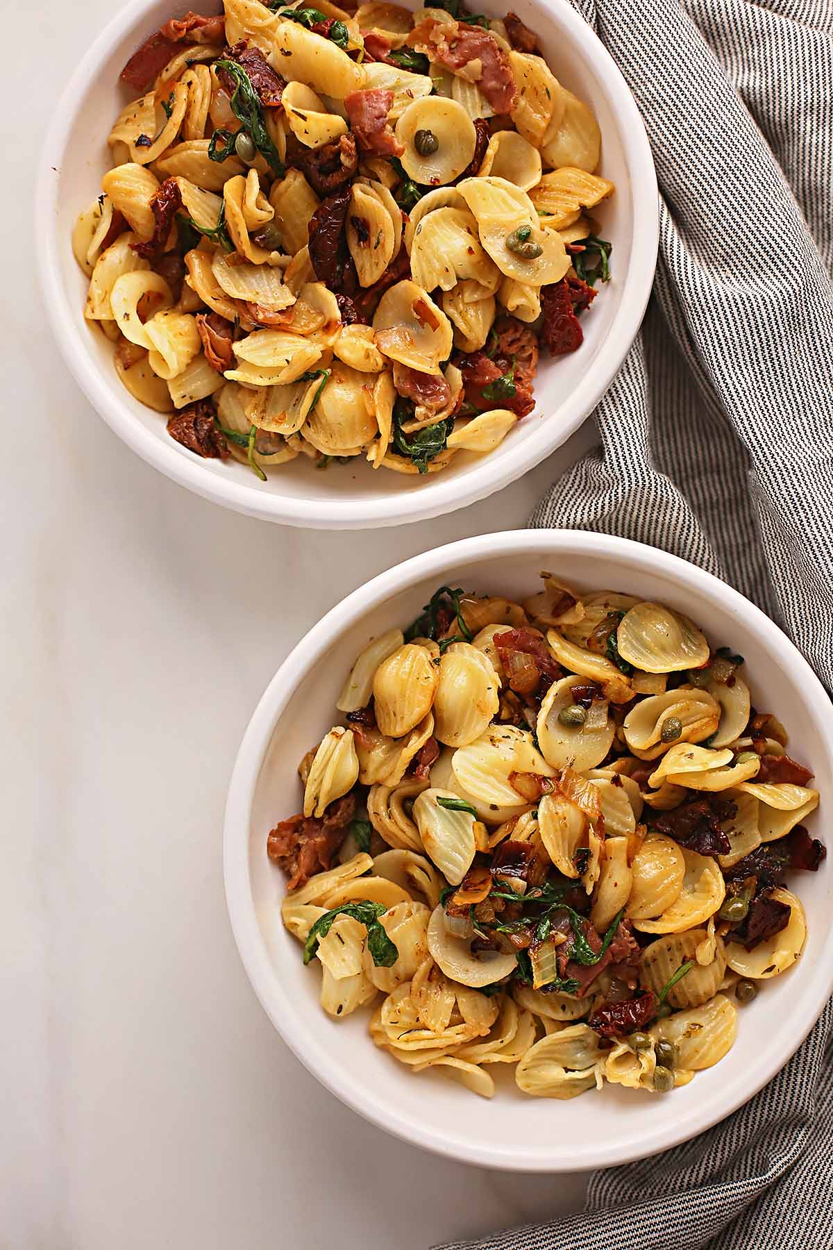 two white bowls served with pasta shells, capers and cooked prosciutto pieces 