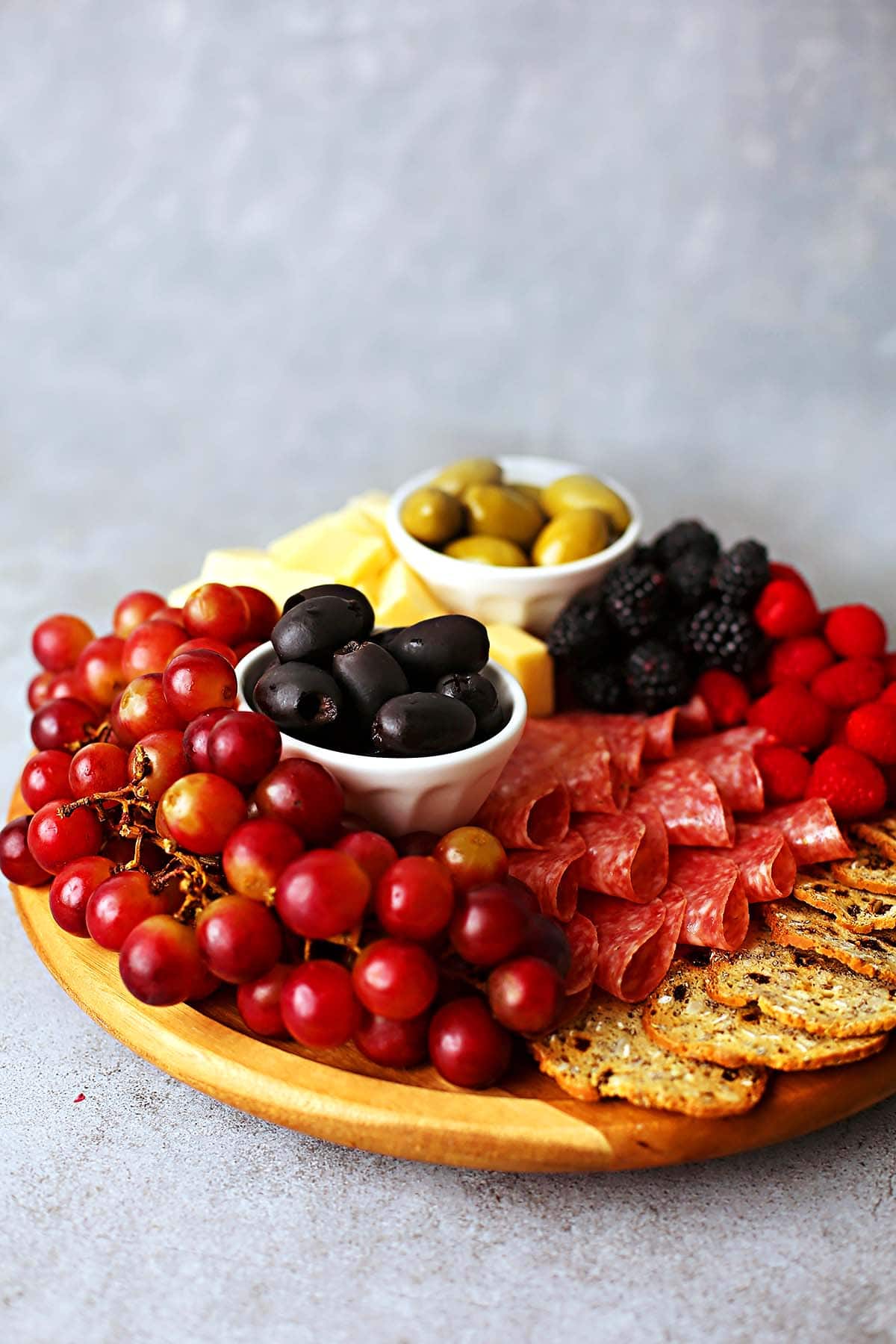 small wooden charcuterie board filled with colorful fruits, crackers, olives.
