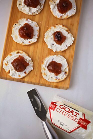 small log of goat cheese next to cutting board served with crackers topped with white cheese and jam