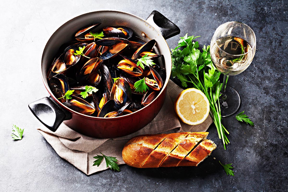 Small pot filled with cooked mussels topped with green herbs and bread next to it. 
