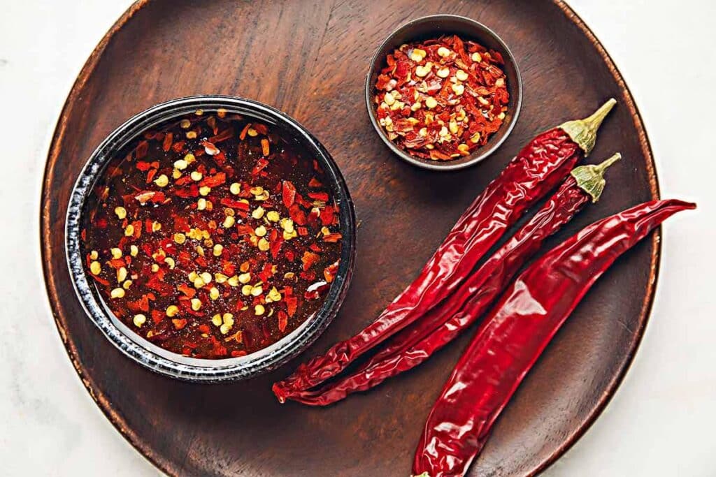 Two bowls with red pepper flakes and red chili peppers next to it. 