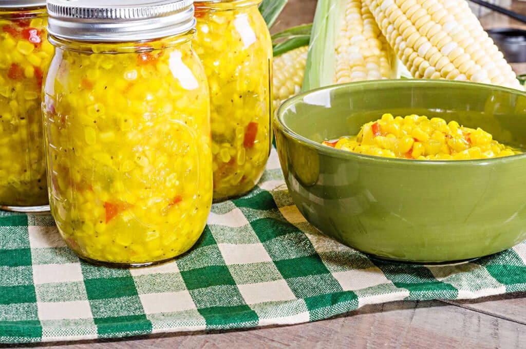 Green bowl with corn relish and glass jar filled with corn next to it.