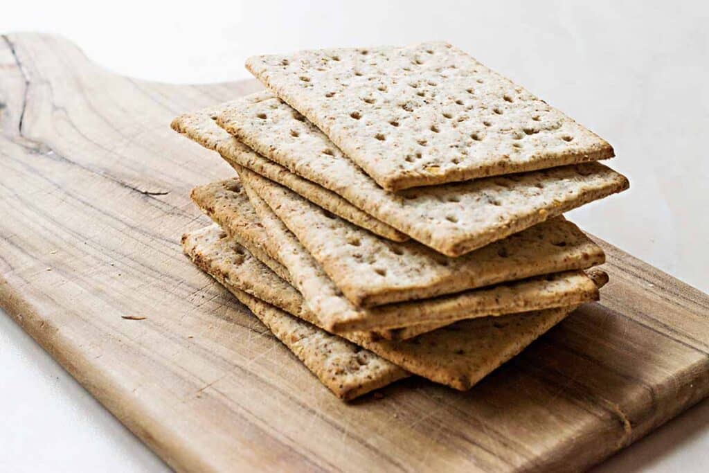 Stack of whole wheat crackers on top of wooden cutting board. 