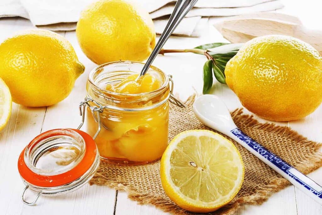 Glass jar filled with lemon preserves next to freshly sliced fruits. 