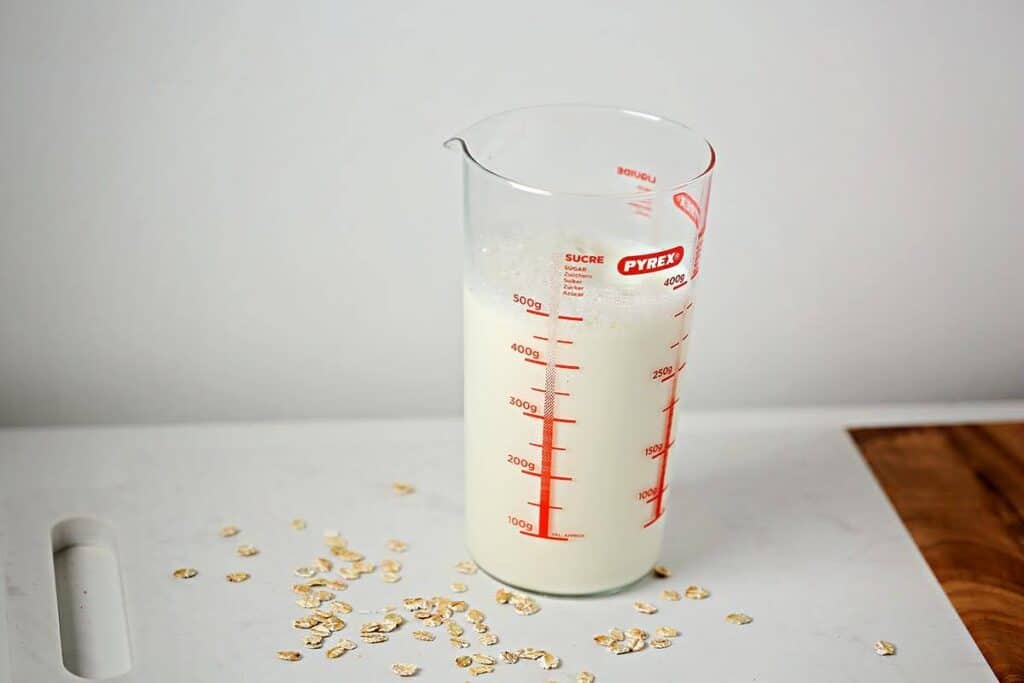 Measuring cup filled with oat milk with oats laying next to it. 
